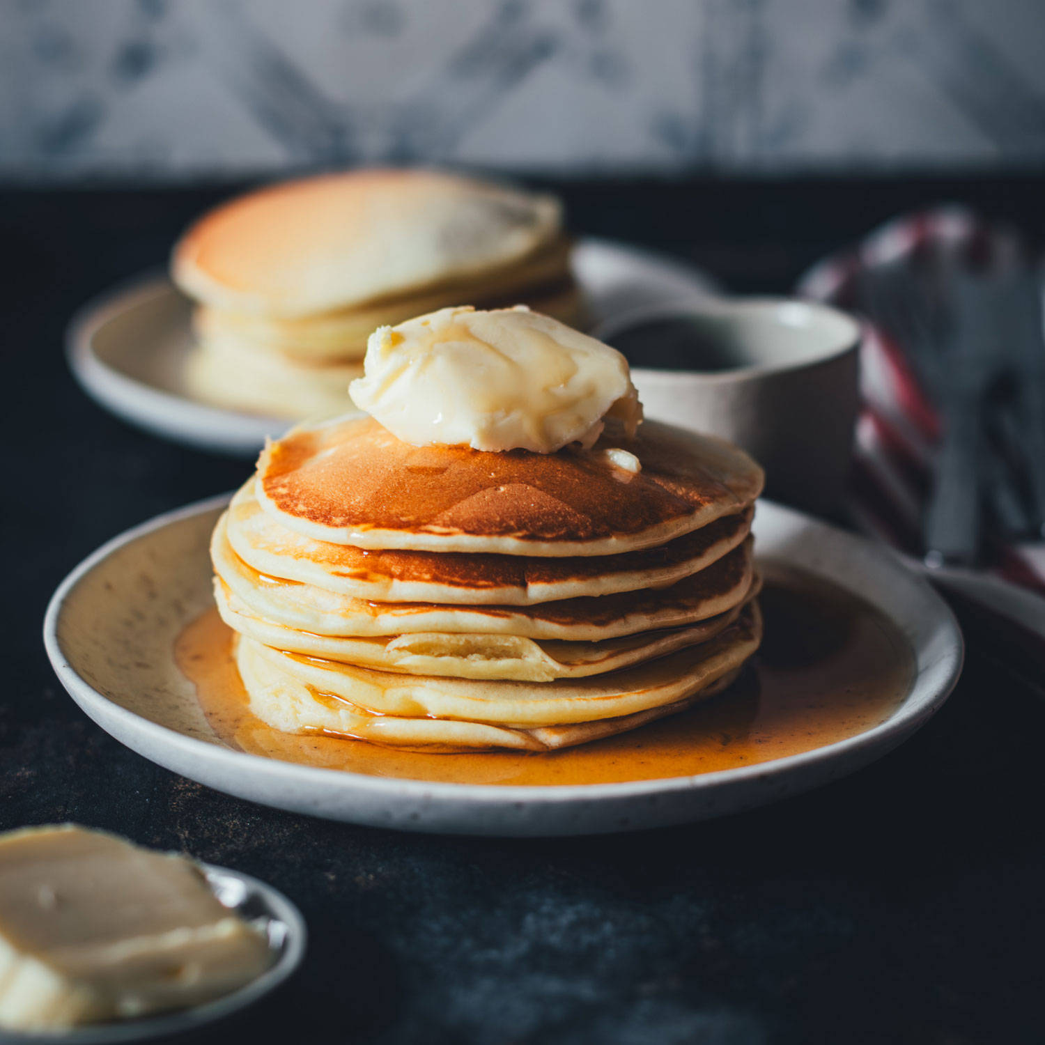 Ein Stapel Pancakes, seitlich fotografiert als quadratisches Bikd. 6 Pancakes auf einem kleinen Teller, übereinander gestapelt und mit einem Stück Salzbutter und Ahornsirup serviert | moeyskitchen.com