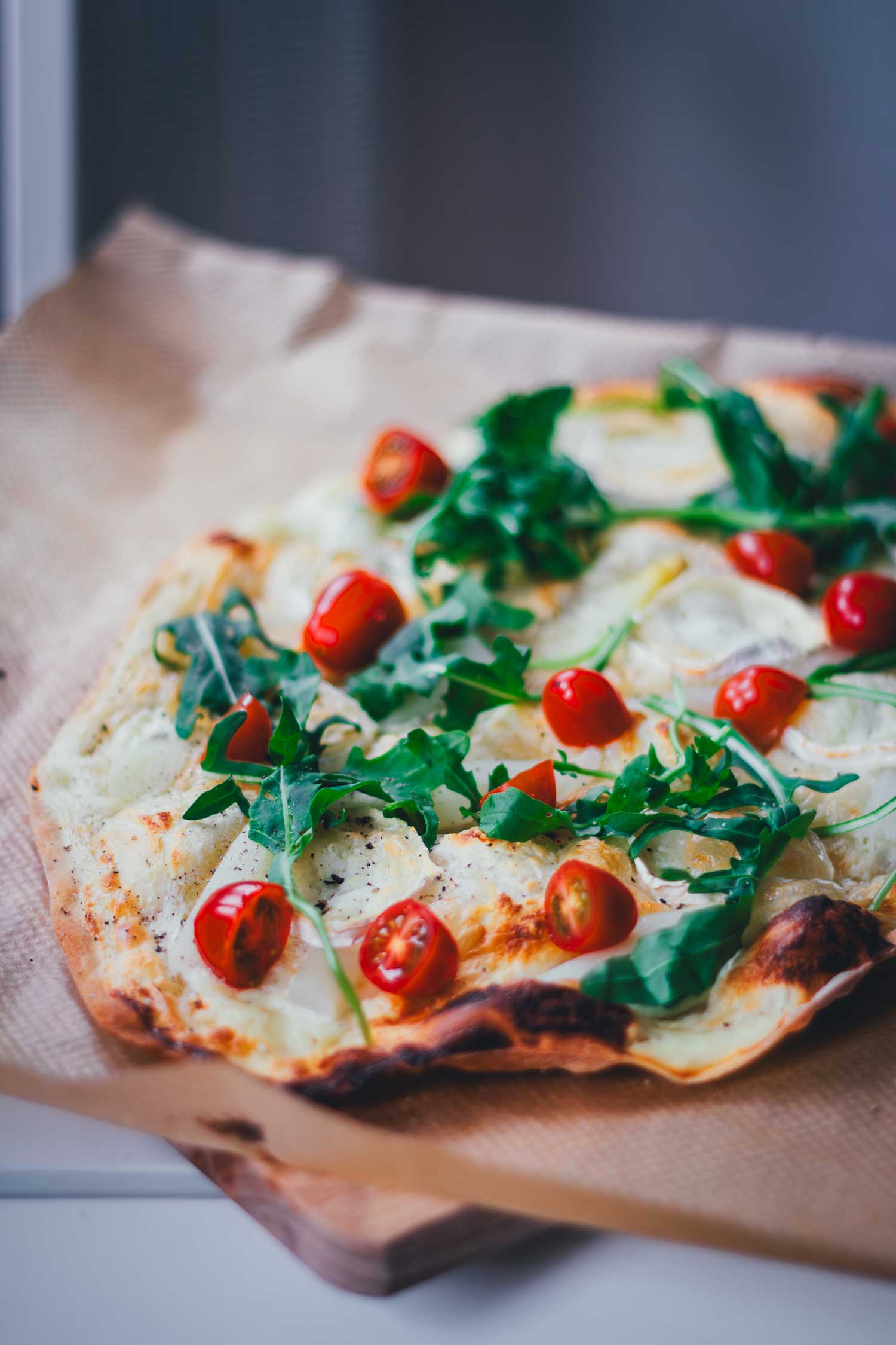 Nahaufnahme im Anschnitt: Spargel-Flammkuchen mit Ziegenkäse und Rucola, serviert auf einem Holzbrett mit Backpapier.
