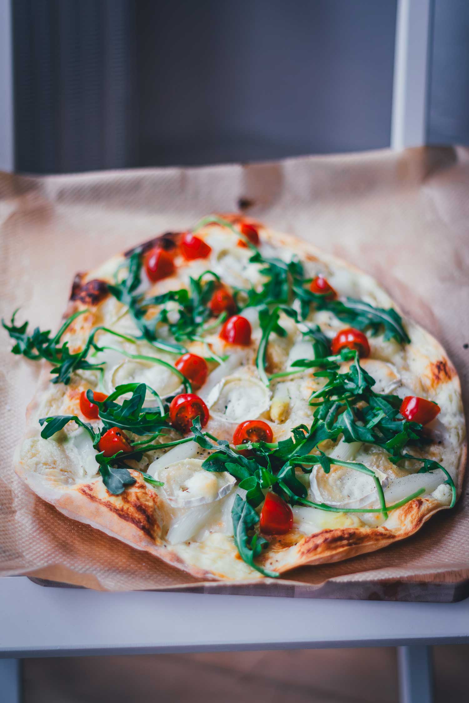 Frontale Ansicht auf einen knusprugen Flammkuchen, der mit weißem Spargel, halbierten Cocktailtomaten, Rucola und Ziegenkäserolle belegt ist. Auf einem Backpapier auf enem Holzbrett servierbereit.