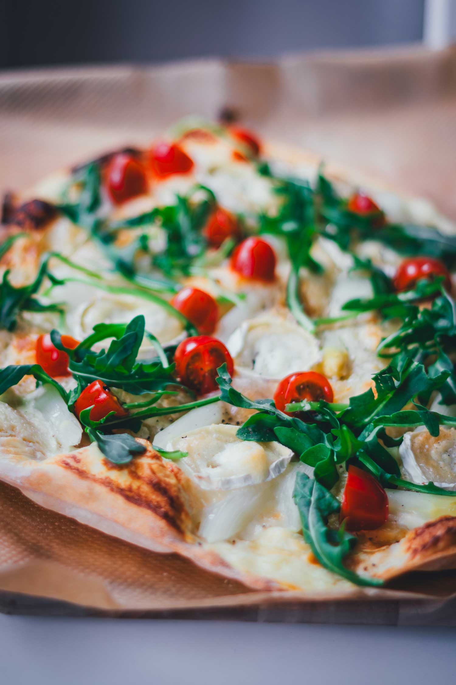 Flache Nahaufnahme von einem frisch gebackenen, knusprigen Flammkuchen  mit weißem Spargel, Tomaten, Rucola und Ziegenkäse.