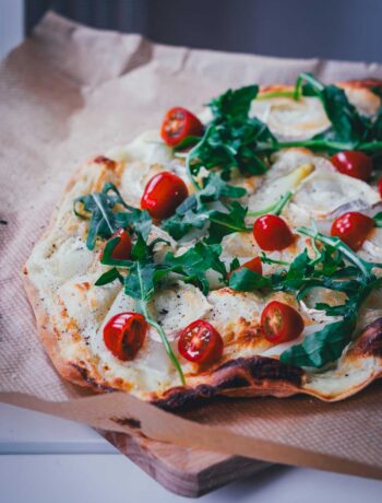 Seitliche Ansicht auf einen frisch gebackenen, knusprigen Flammkuchen mit Spargel. Zusätzlich belegt mit kleinen Tomaten, Rucola und Ziegenkäserolle perfekt für den Frühling!