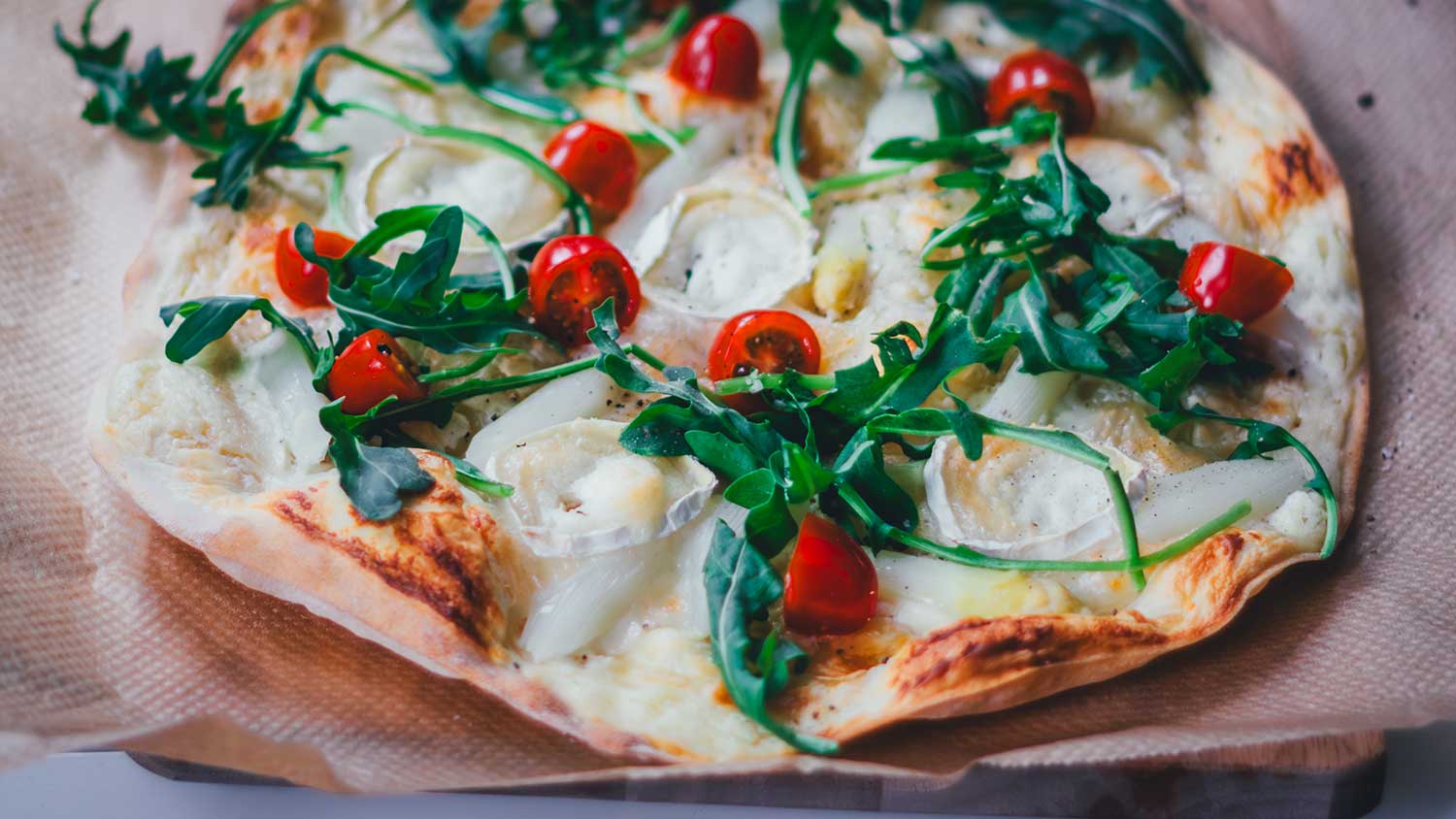 Nahaufnahme von einem knusprigen Spargel-Flammkuchen, der mit weißem Spargel, Rucola, Tomaten und Ziegenkäse belegt ist.