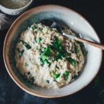 Leicht seitliche Nahaufnahme von einem tiefen Teller, gefüllt mit cremigem Bärlauch Risotto mit Erbsen und Frühlingszwiebeln. Daneben liegen eine Serviette und eine Schale mit geriebenem Käse.