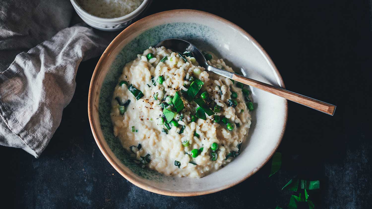Leicht seitliche Nahaufnahme von einem tiefen Teller, gefüllt mit cremigem Bärlauch Risotto mit Erbsen und Frühlingszwiebeln. Daneben liegen eine Serviette und eine Schale mit geriebenem Käse.