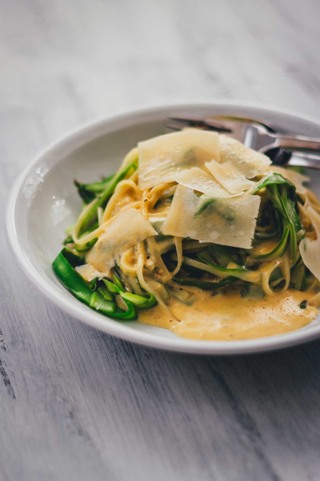 Pasta mit grünem Spargel und Parmesansauce auf einem Teller mit Parmesanspänen serviert, seitlich zu sehen.