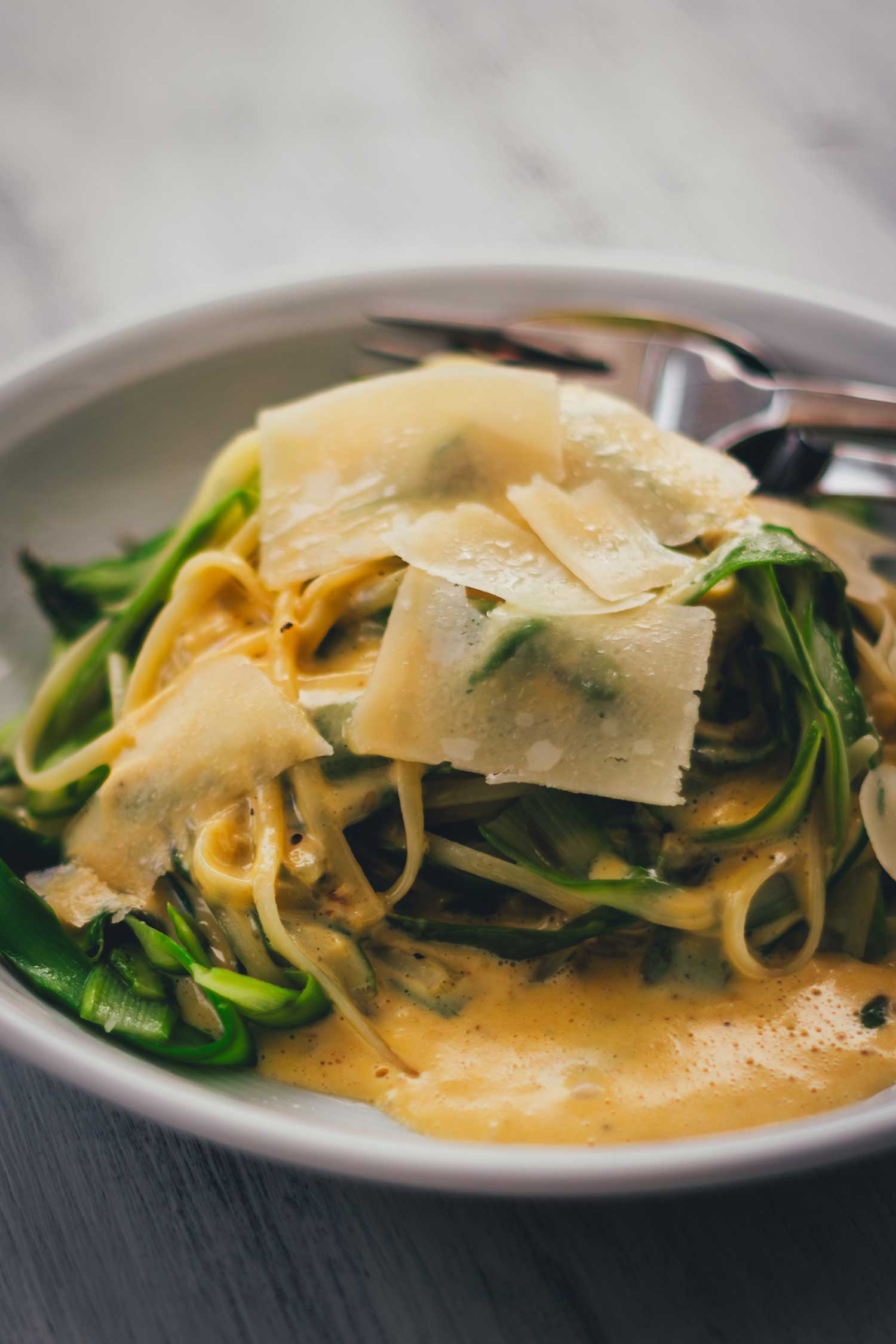 Nahaufnahme von einem Teller mit frischer Pasta mit grünem Spargel und cremiger Parmesansauce, serviert mit Parmesanspänen.