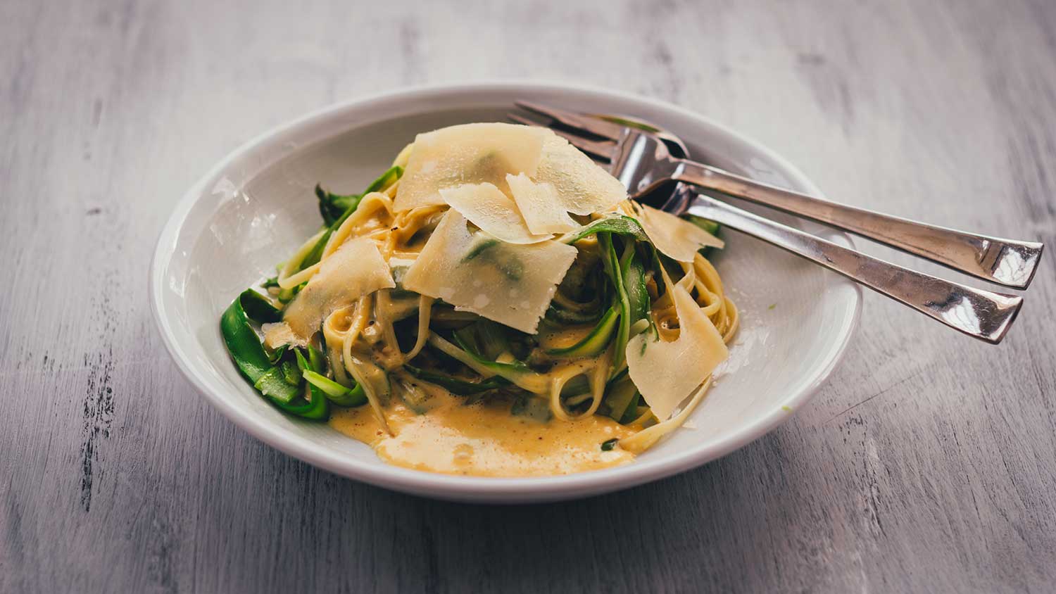 Pasta mit grünem Spargel und creiger Parmesansauce auf einem weißen Teller, garniert mit Parmesanspänen und frisch gemahlenem Pfeffer