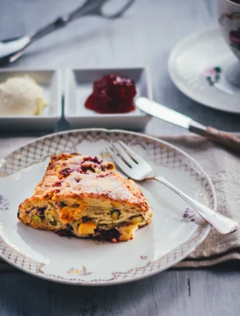 Himbeer Scones mit Pistazien und weißer Schokolade auf einem Goldrand-Teller, dazu Clotted Cream und Himbeerkonfitüre in kleinen Schälchen