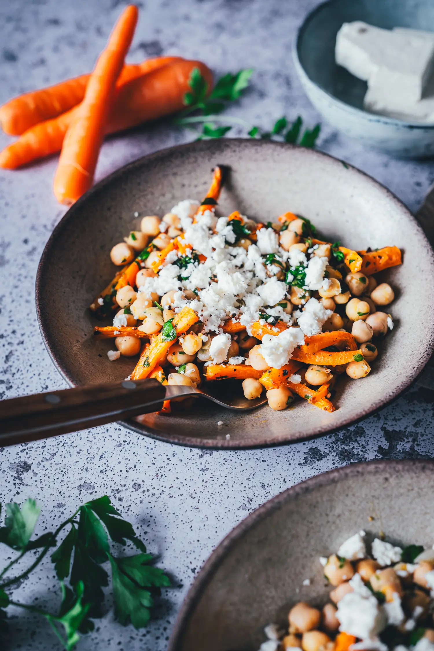 Nahaufnahme einer Schale Kichererbsen-Salat mit gerösteten Möhrenstiften, Feta und frischer Petersilie, daneben liegen rohe Möhren