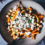 Schale Kichererbsen-Salat mit gerösteten Möhren, Feta und frischer Petersilie mit Holzgabel, umgeben von rohen Möhren und Feta