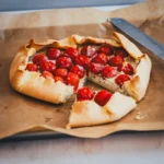 Tomaten-Galette mit Ricotta und ganzen Kirschtomaten, aufgeschnitten auf einem mit Backpapier ausgelegten Blech
