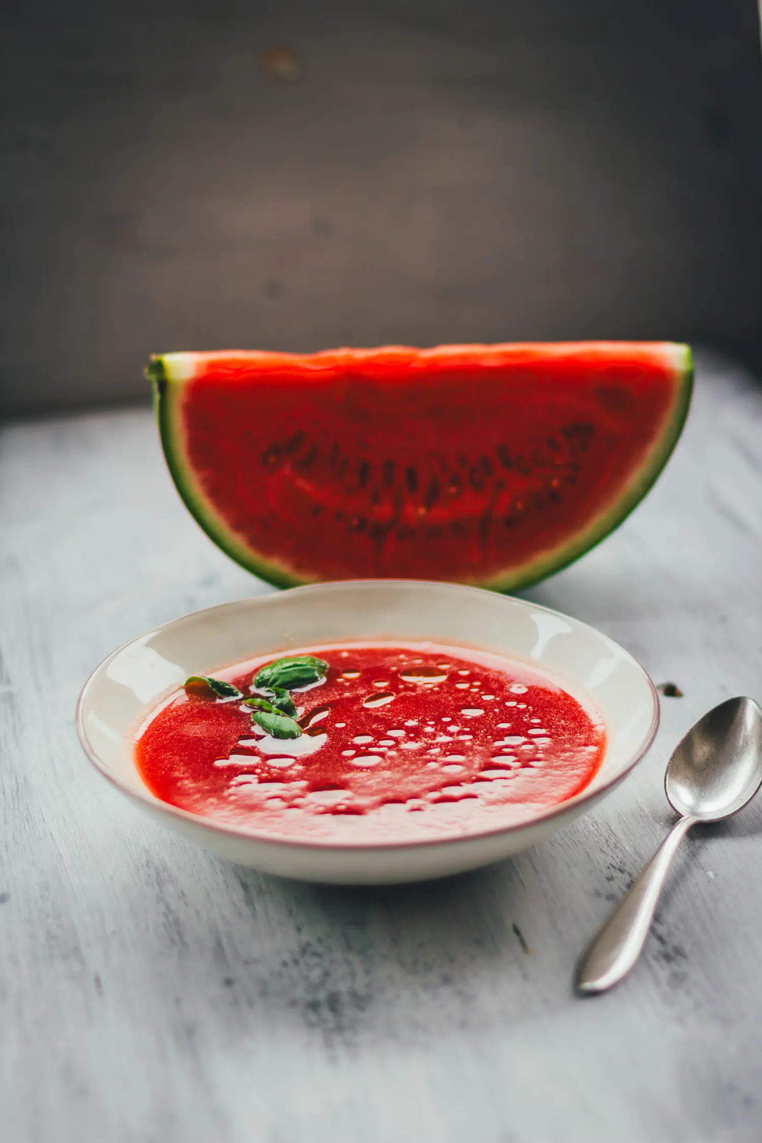 Kalte Sommersuppe aus Wassermelone in einer Schale, im Hintergrund ein Stück Wassermelone und ein Löffel