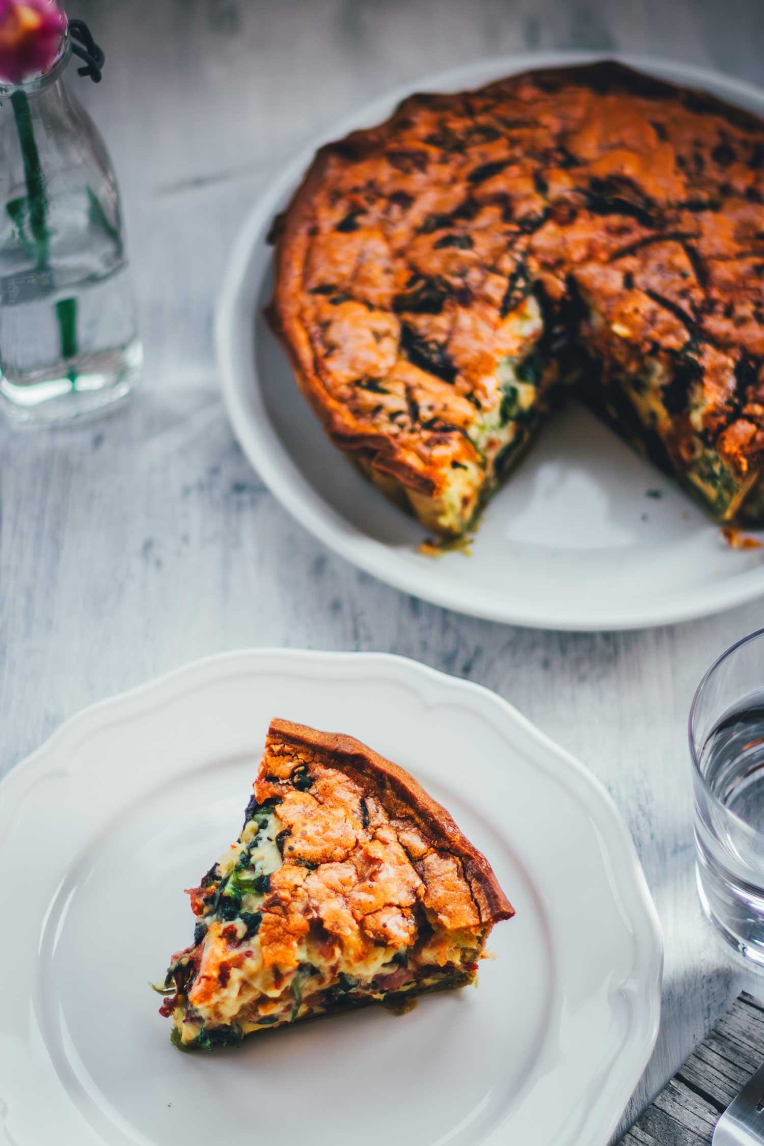 Deatialreiche Nahaufnahme von schräg oben auf ein Stück Spinat-Bacon-Quiche auf einem weißen Teller. Dahinter sieht man die angeschnittene Quiche liegen.