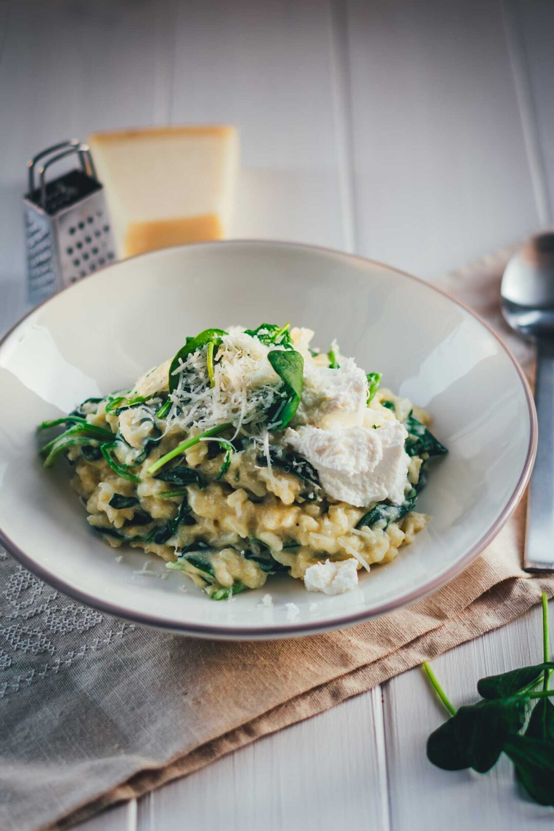Man sieht einen hellgrauen Teller, der mit cremigem Ofenrisotto mit Spinat gefüllt ist. Daneben liegen Löffel, Parmesan und Spinat. Das Gericht sieht sehr cremig aus und wir mit Käse getoppt, bereit zum Servieren.
