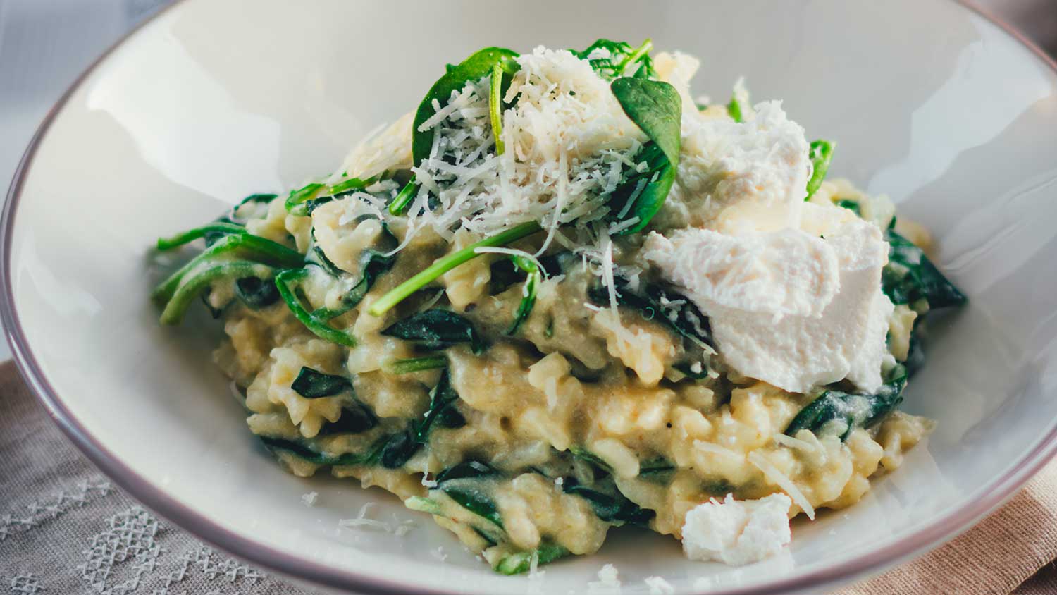 Detaillierte Nahaufnahme eines tiefen Tellers, gefüllt mit cremigem Ofenrisotto mit Spinat. Mit Ricotta und Parmesan bestreut serviert, sieht man, wie cremig und aroamtisch das Gericht ist.