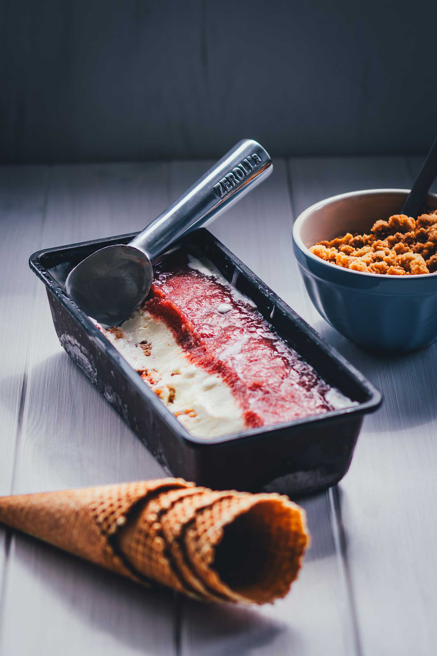 Rhabarber-Eis in der Eisform mit Eisportionierer, daneben eine Schüssel mit knusprigen Streuseln zum Bestreuen.