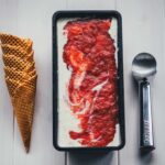 Selbstgemachtes Rhabarber-Eis mit Streuseln in einer schwarzen Eisform, daneben gestapelte Waffelhörnchen und ein Eisportionierer.
