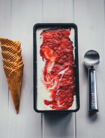 Selbstgemachtes Rhabarber-Eis mit Streuseln in einer schwarzen Eisform, daneben gestapelte Waffelhörnchen und ein Eisportionierer.