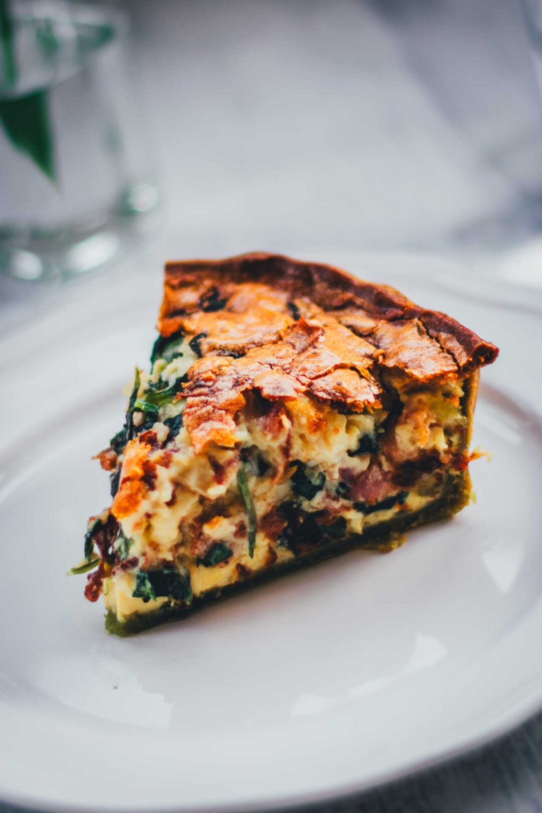 Nahaufnahme von einem einzelnen Stück Spinat-Bacon-Quiche auf einem weißen Teller. Man erkennt den grünen Spinat-Ricotta-Boden und die saftige Füllung der Quiche.
