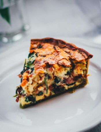 Nahaufnahme von einem einzelnen Stück Spinat-Bacon-Quiche auf einem weißen Teller. Man erkennt den grünen Spinat-Ricotta-Boden und die saftige Füllung der Quiche.