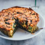 Spinat-Bacon-Quiche auf einem Teller serviert. Ein Stück wurde bereits herausgeschnitten und man erkennt den grünen Ricotta-Spinat-Boden und die würzige Füllung mit Bacon und Spinat.