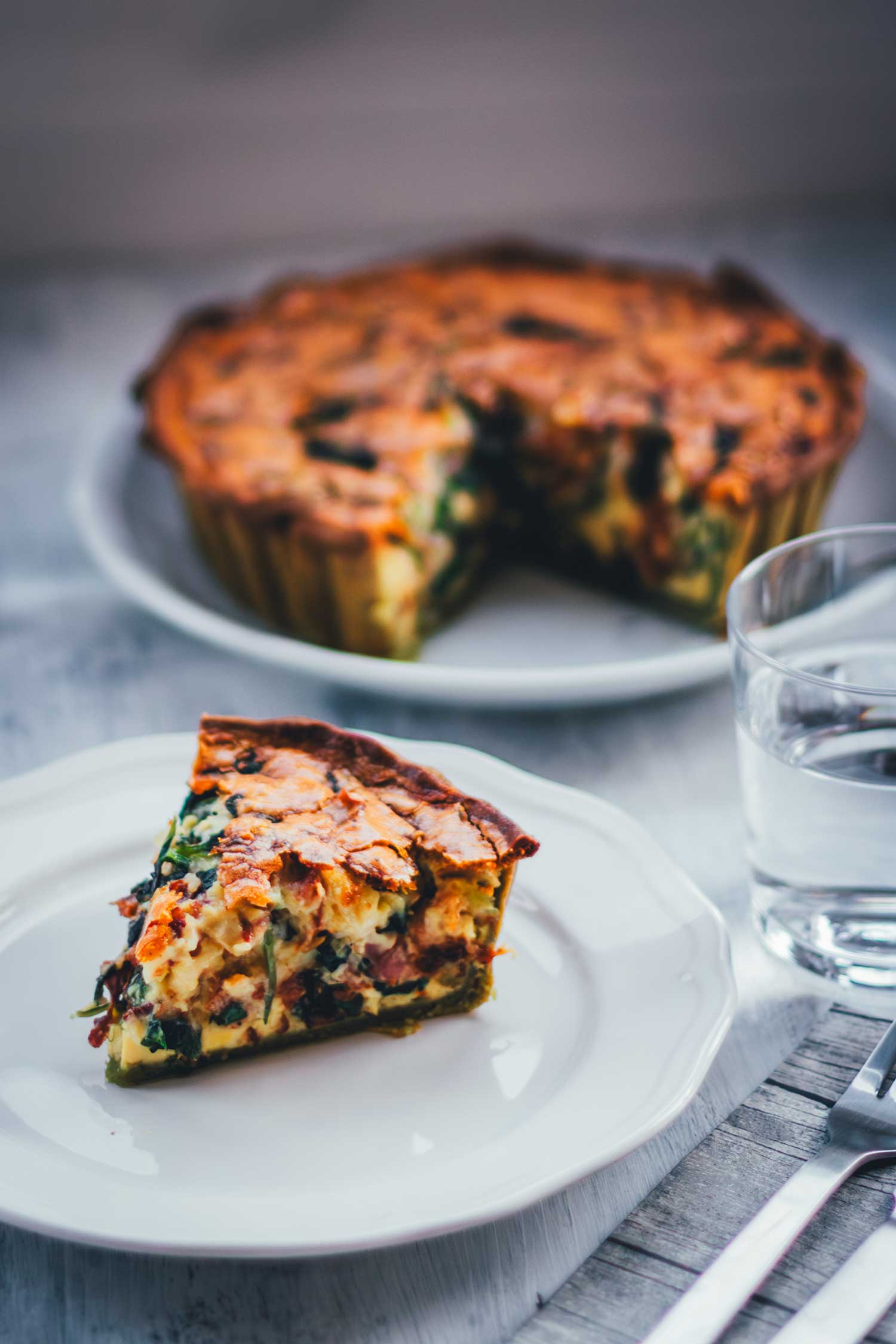 Seitliche Ansicht von einem Stück Quiche auf einem weißen Teller, daneben liegen Serviette und Besteck sowie ein Glas Wasser. Im Hintergrund sieht man die angeschnittene Quiche stehen.