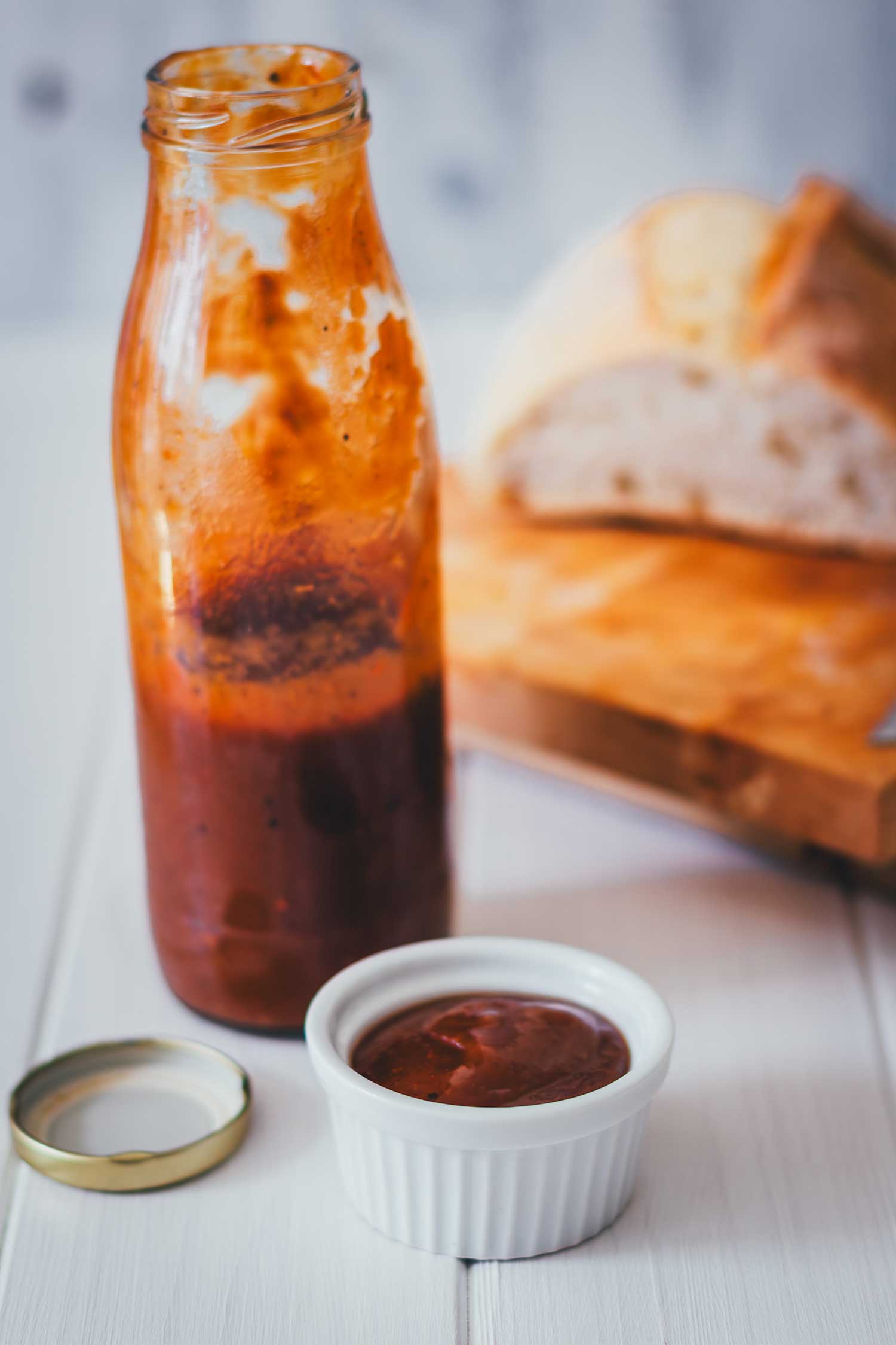 Glasflasche mit Rhabarber-Ketchup und gefülltes Schälchen, im Hintergrund aufgeschnittenes Brot auf einem Holzbrett