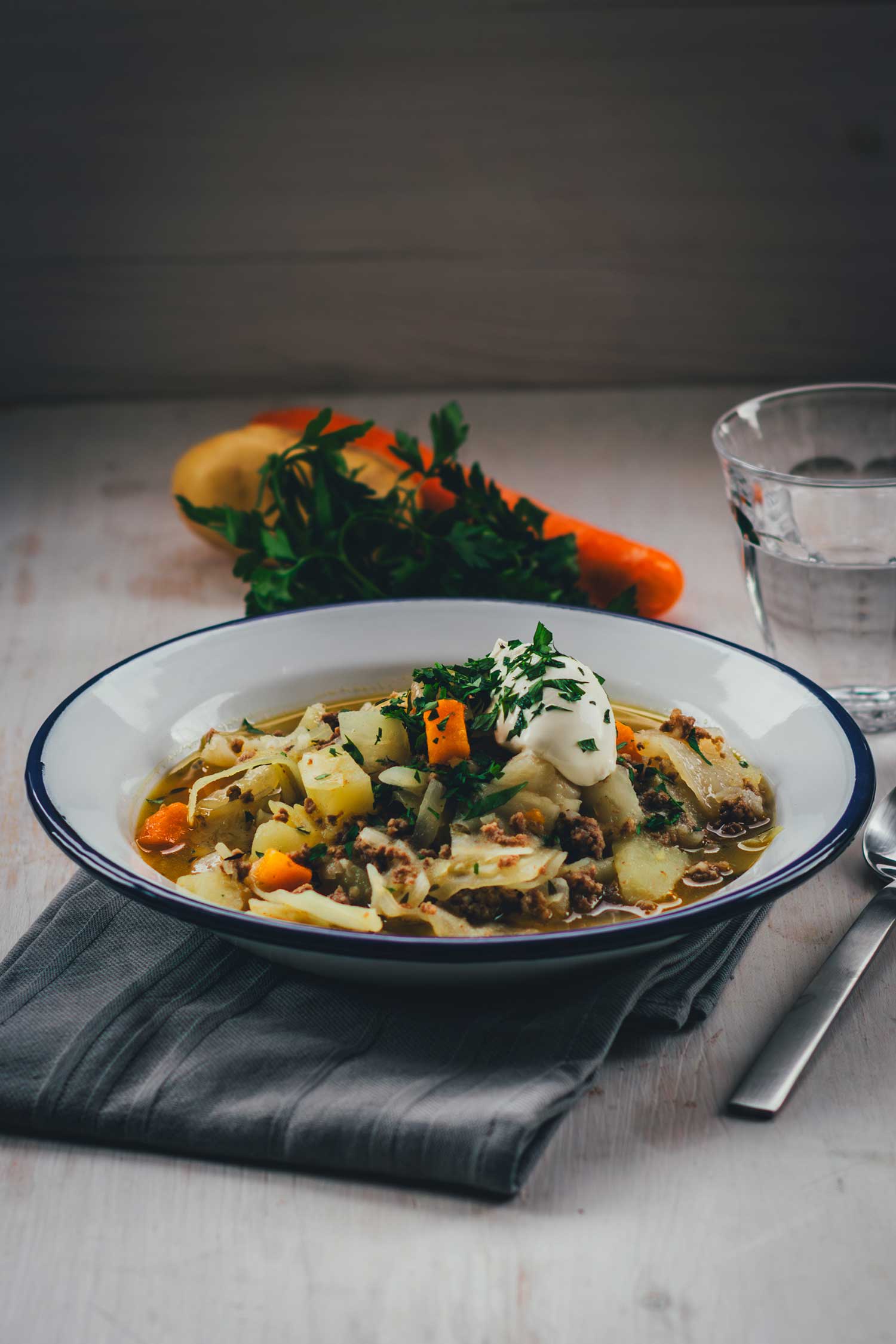 seitliche Ansicht einer Essenszene: Eintopf in einem tiefen Teller serviert, drumherum Gemüse, ein Glas Wasser und ein Esslöffel | moeyskitchen.com