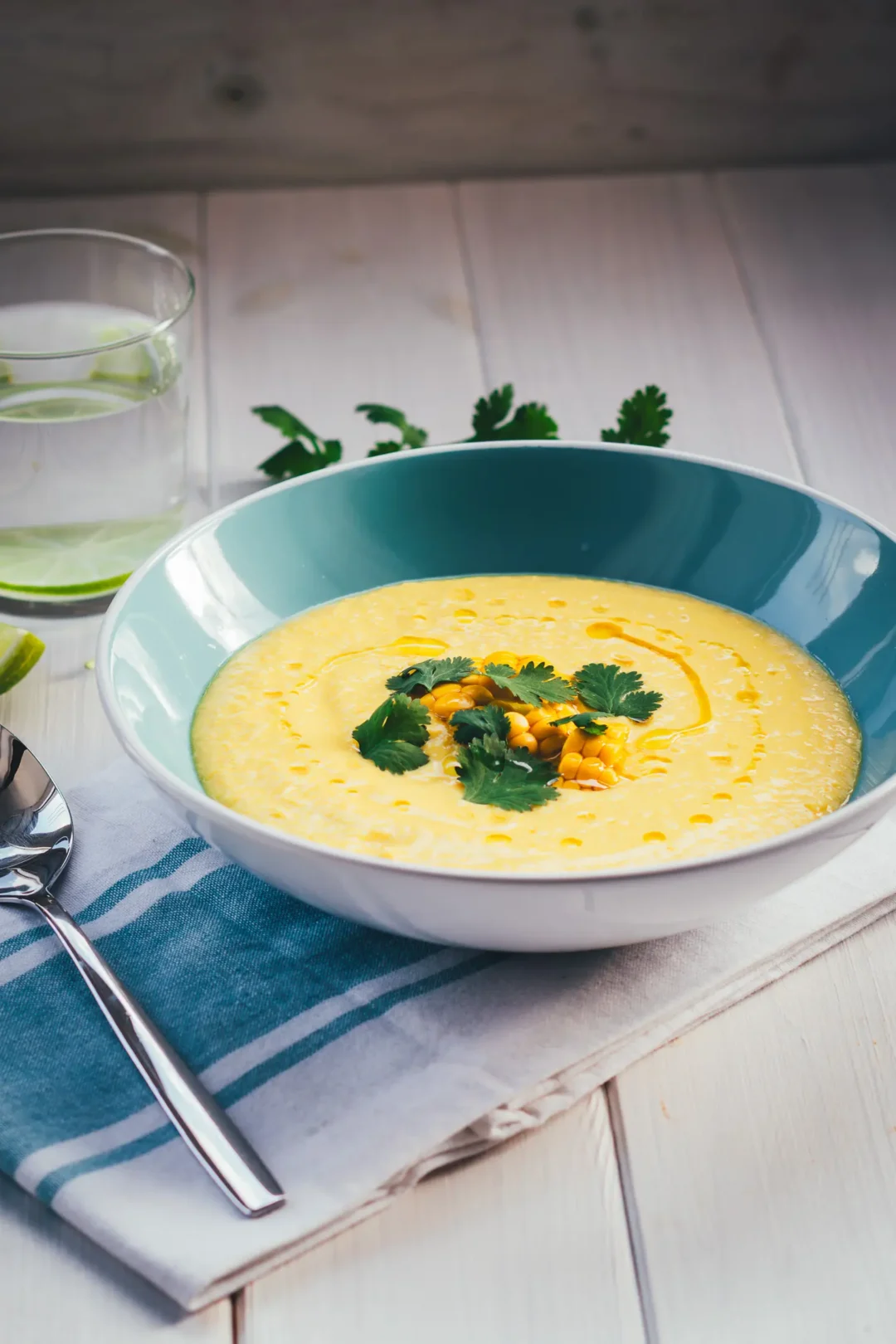 Blaue Schüssel mit gelber Mais-Paprika-Gazpacho, die mit Koriander getoppt ist. Daneben liegt eine blau gestreifte Serviette mit einem Löffel, im Hintergrund steht ein Wasserglas mit Limettenscheibe.