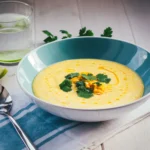 Blaue Schüssel mit gelber Mais-Paprika-Gazpacho, die mit Koriander getoppt ist. Daneben liegt eine blau gestreifte Serviette mit einem Löffel, im Hintergrund steht ein Wasserglas mit Limettenscheibe.