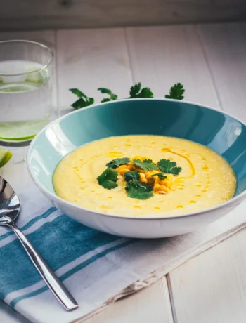 Blaue Schüssel mit gelber Mais-Paprika-Gazpacho, die mit Koriander getoppt ist. Daneben liegt eine blau gestreifte Serviette mit einem Löffel, im Hintergrund steht ein Wasserglas mit Limettenscheibe.