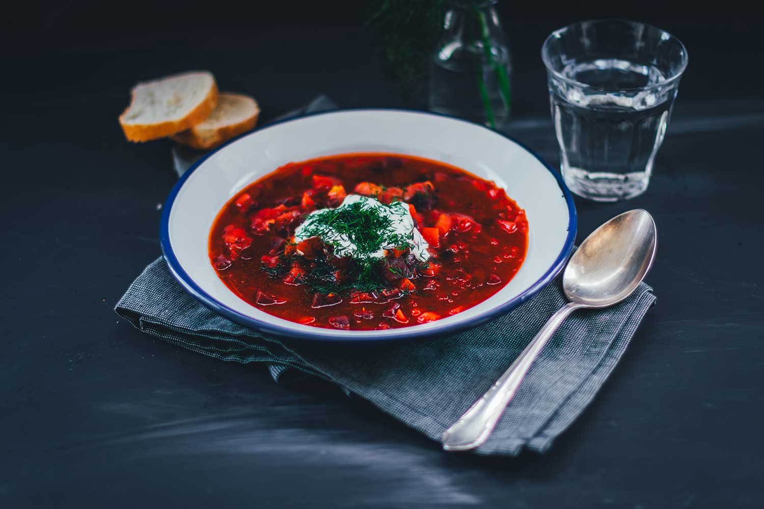 Querformatiges Stillleben von einem Teller mit rotem Eintopf, daneben ein Löffel, darunter euine Serviette. Man sieht Brot drumherum und ein Glas Wasser, sowie etwas Dill. | moeyskitchen.com