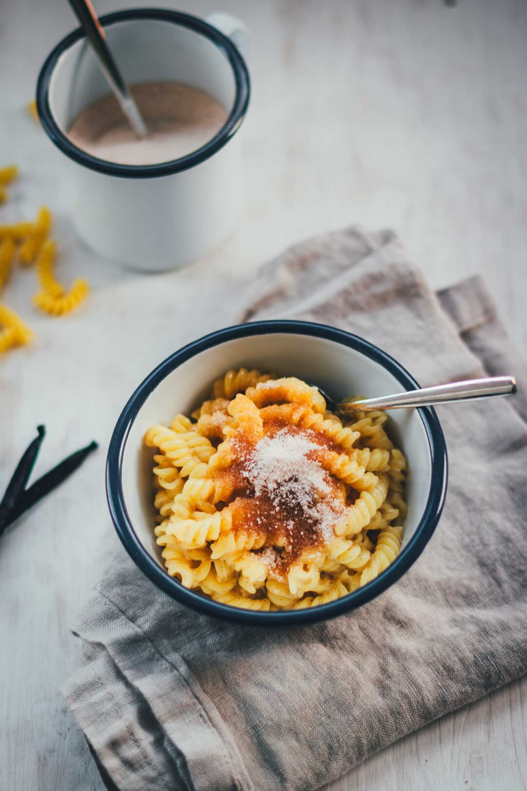 Eine Schüssel cremige Milchnudeln, zubereitet nach Omas Rezept mit Sahne und braunem Zucker, bestreut mit Zimt-Zucker und angerichtet auf einem grauen Leinentuch. Im Hintergrund sind Vanilleschoten, rohe Nudeln und Zimt und Zucker zu sehen.