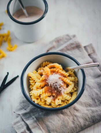 Eine Schüssel cremige Milchnudeln, zubereitet nach Omas Rezept mit Sahne und braunem Zucker, bestreut mit Zimt-Zucker und angerichtet auf einem grauen Leinentuch. Im Hintergrund sind Vanilleschoten, rohe Nudeln und Zimt und Zucker zu sehen.