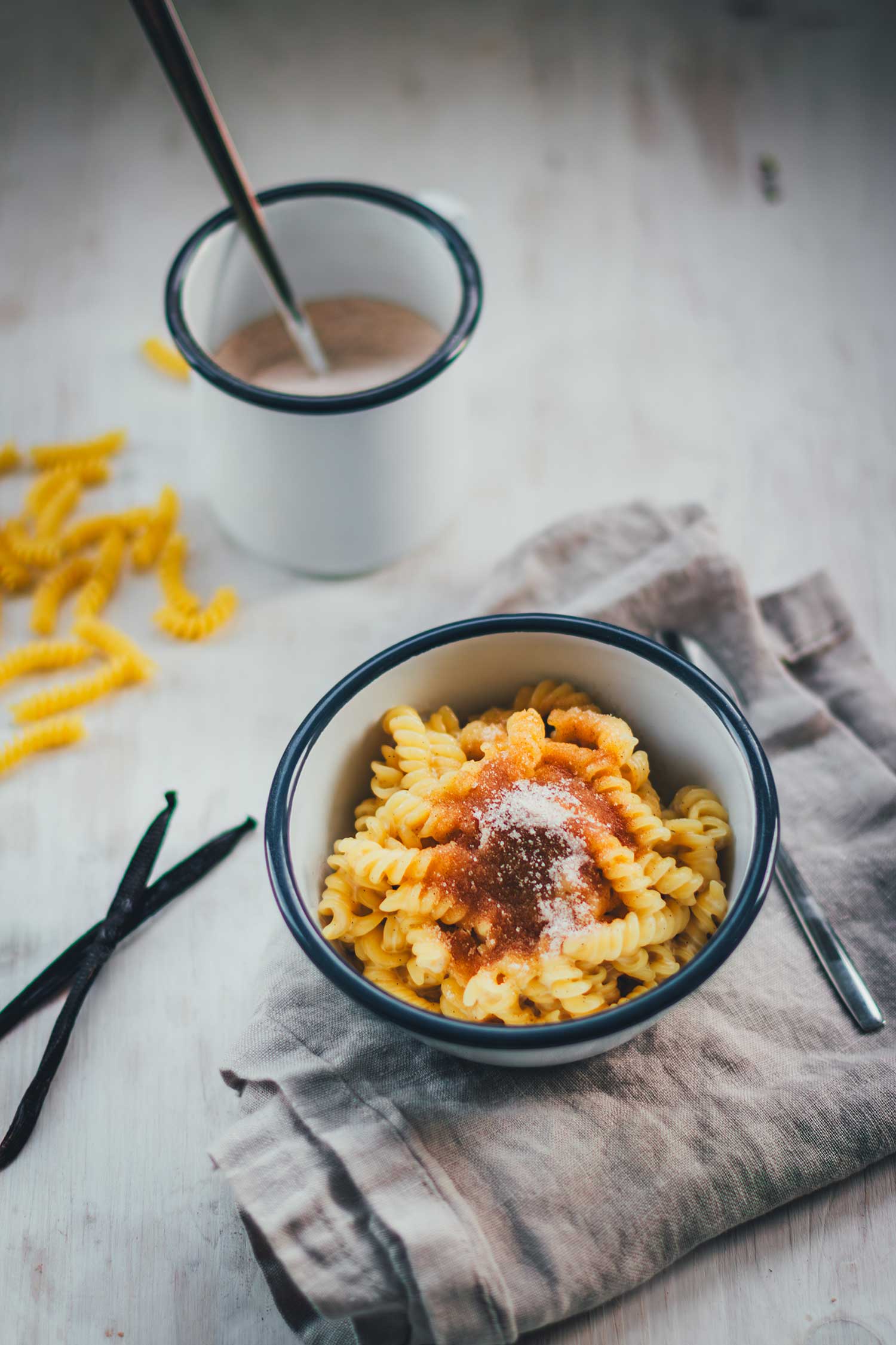 Eine Schrägansicht, die die Schüssel mit süßer Pasta auf dem grauen Leinentuch zeigt. Im Hintergrund sind Vanilleschoten, Roh-Nudeln und die Zimt-Zucker-Mischung in einem Becher zu sehen.
