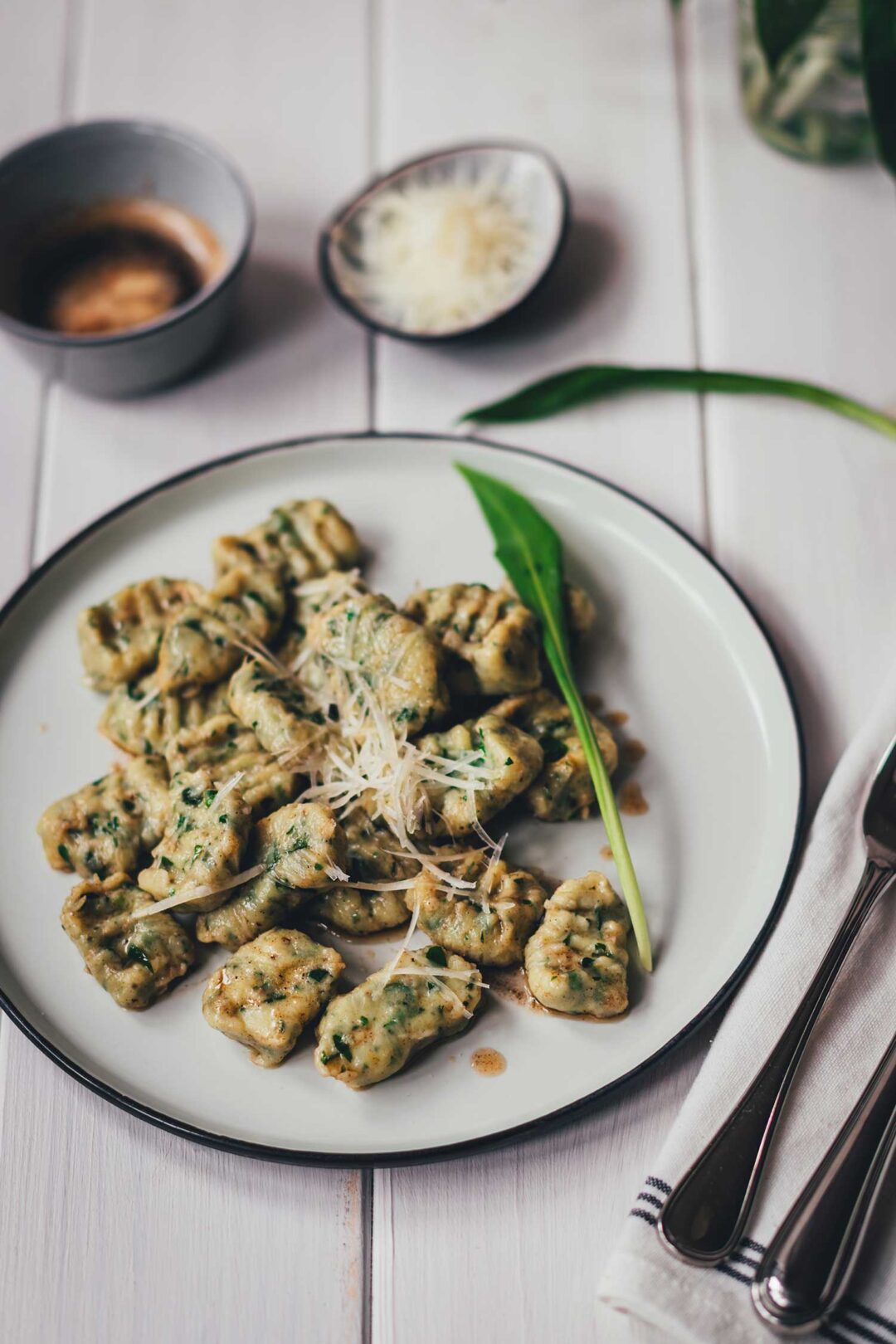 Seitliche, leicht schräge Draufsicht auf einen Teller mit frisch gekochten Bärlauch-Ricotta-Gnocchi. Serviert mit brauner Butter und geriebenem Käse im Hintergrund.