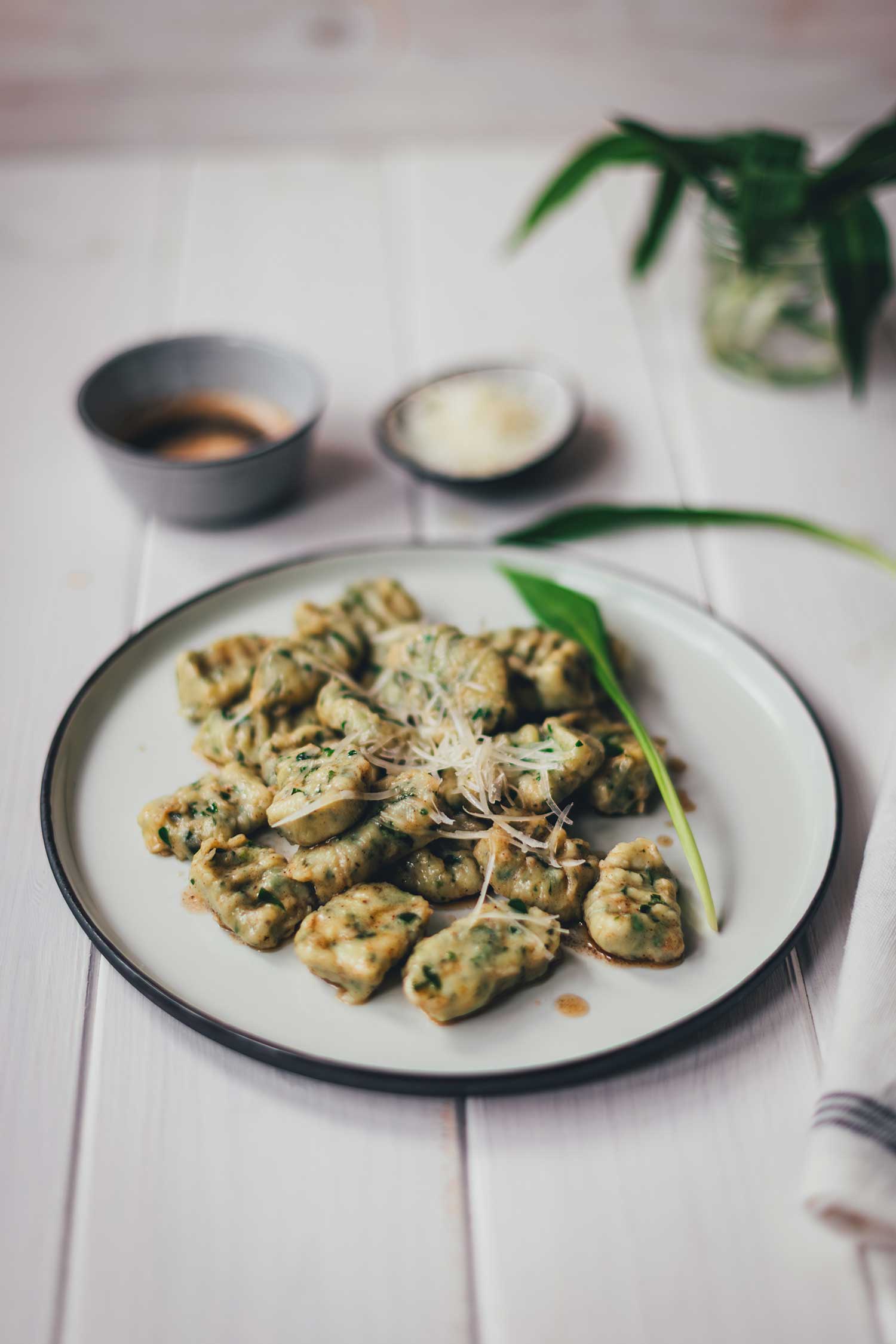 Ein heller Teller mit selbst gemachten Bärlauch-Rciotta-Gnocchi, servierbereit. Die Gnocchi sind in Butter geschwenkt und mit Käse bestreut. Beides sieht man im Hintergrund mit etwas frischem Bärlauch.