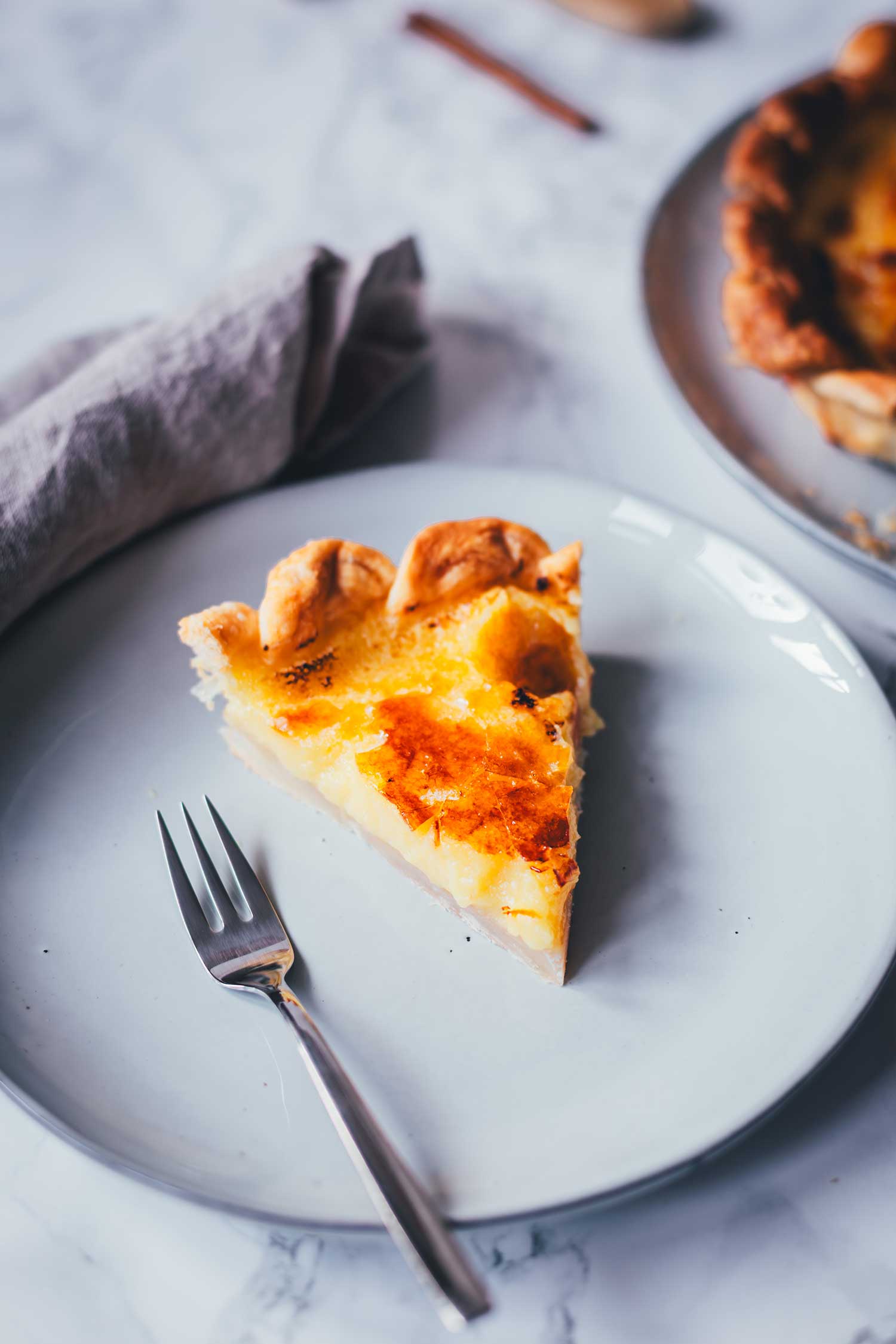 Nahaufnahme eines einzelnen Kuchenstücks einer Pasteis de Nata Tarte auf einem hellgrauen Teller mit einer Kuchengabel. Im Hintergrund liegt eine graue Serviette.
