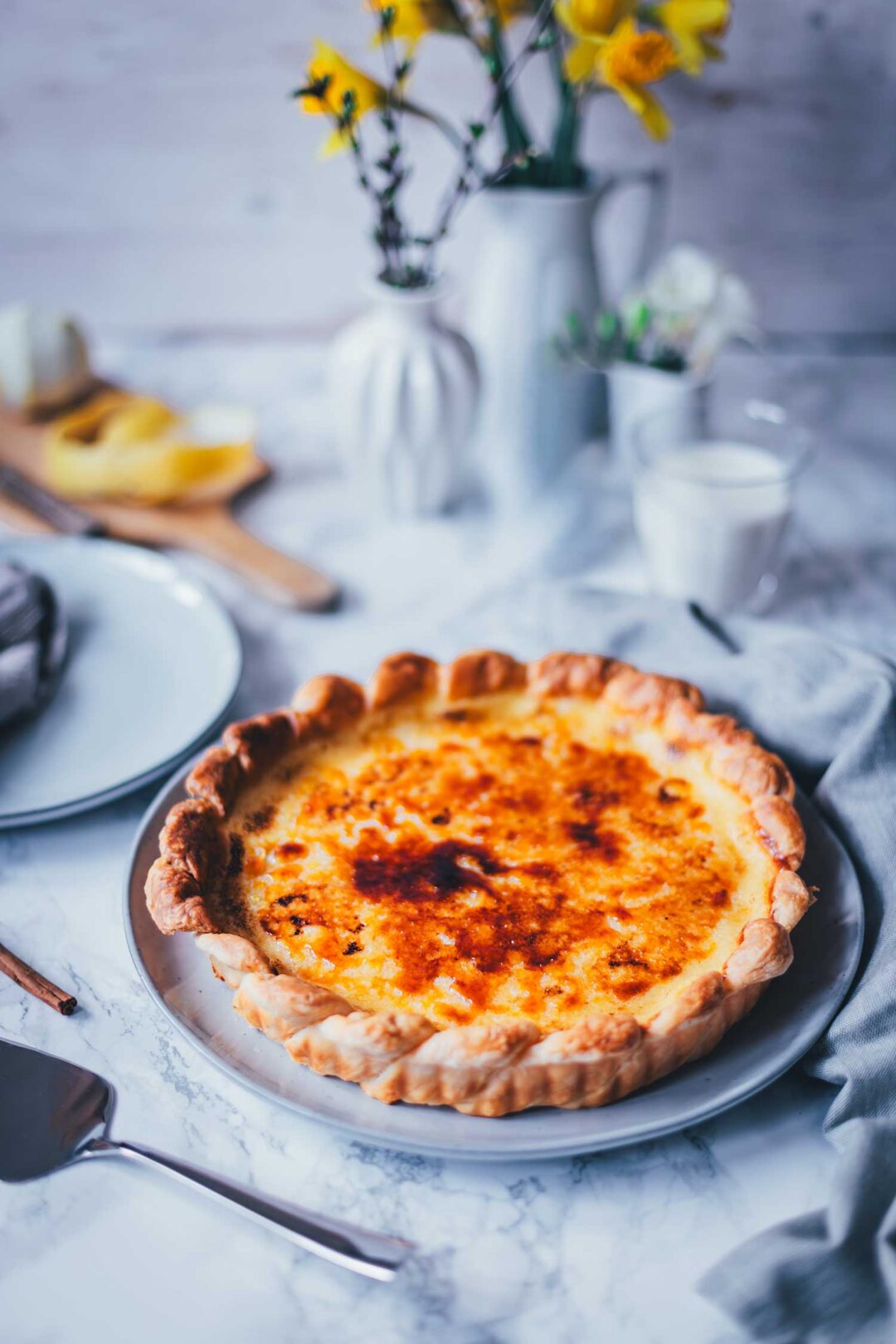 Seitliche Ansicht von einer großen Pasteis de Nata tarte auf einem runden Teller. Man erkennt die goldgelbe Füllung, den karamellisierten Zucker und den knusprigen Blätterteig. Im Hintergrund sieht man gelbe Blumen in weißen Vasen und einen marmorfarbenen Untergrund.
