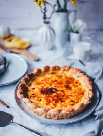 Seitliche Ansicht von einer großen Pasteis de Nata tarte auf einem runden Teller. Man erkennt die goldgelbe Füllung, den karamellisierten Zucker und den knusprigen Blätterteig. Im Hintergrund sieht man gelbe Blumen in weißen Vasen und einen marmorfarbenen Untergrund.