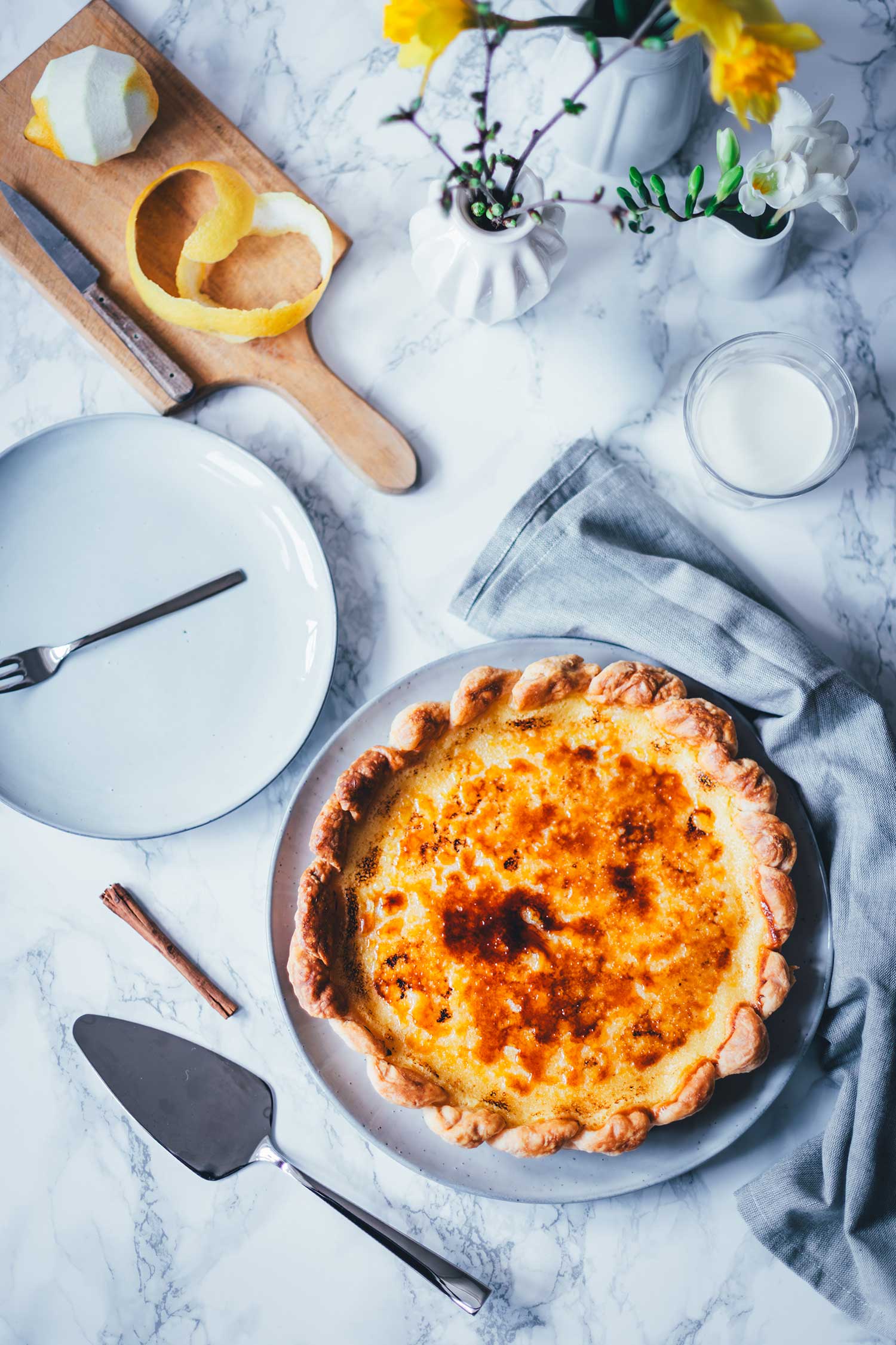 Ansicht von oben eines gedeckten Tischs mit frisch gebackener Pasteis de Nata Tarte auf einem runden Teller, daneben ein Kuchenheber, ein Glas Milch, Zitronenschale, gelbe Blumen in weißer Vase und einen Teller zum Servieren.