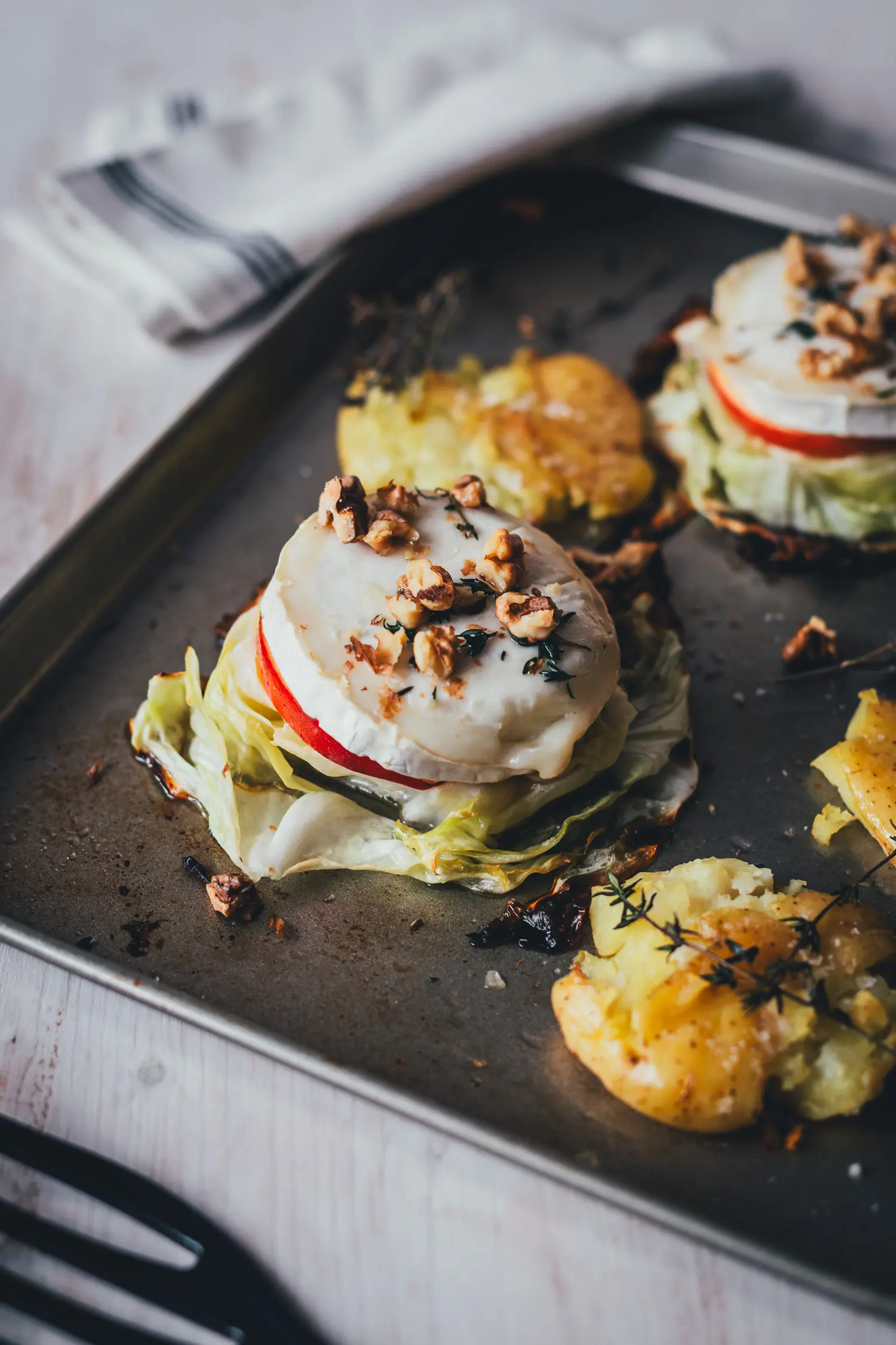 Gebackenes Spitzkohl-Schnitzel aus Spitzkohlscheibe, Apfel und Ziegenkäse mit Walnüssen und Thymian, daneben Smashed Potatoes auf dem Blech.