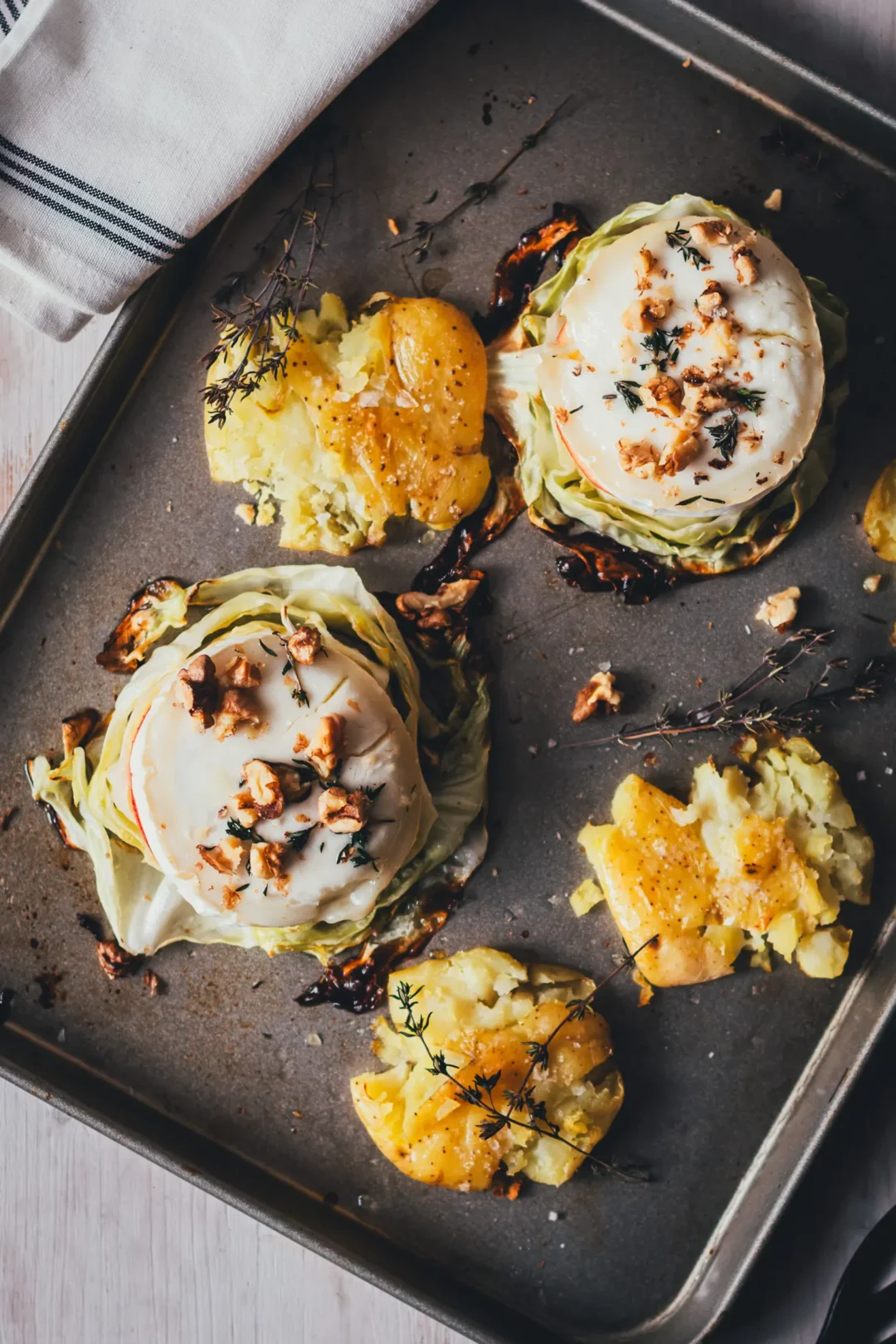 Zwei vegetarische Spitzkohl-Schnitzel mit geschmolzenem Ziegenkäse, Apfelscheibe, Walnüssen und Thymian, daneben knusprige Smashed Potatoes auf einem Backblech.