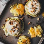 Zwei vegetarische Spitzkohl-Schnitzel mit geschmolzenem Ziegenkäse, Apfelscheibe, Walnüssen und Thymian, daneben knusprige Smashed Potatoes auf einem Backblech.