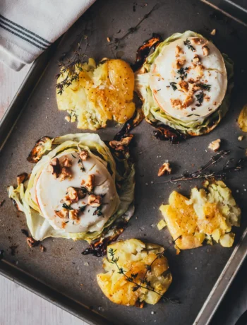 Zwei vegetarische Spitzkohl-Schnitzel mit geschmolzenem Ziegenkäse, Apfelscheibe, Walnüssen und Thymian, daneben knusprige Smashed Potatoes auf einem Backblech.