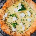 Erbsen-Ricotta-Quiche mit Filoteig in einer schwarzen Tarteform auf einem runden Auskühlgitter, garniert mit Tupfen Ziegenfrischkäse und frischen Minzeblättern, von oben fotografiert.