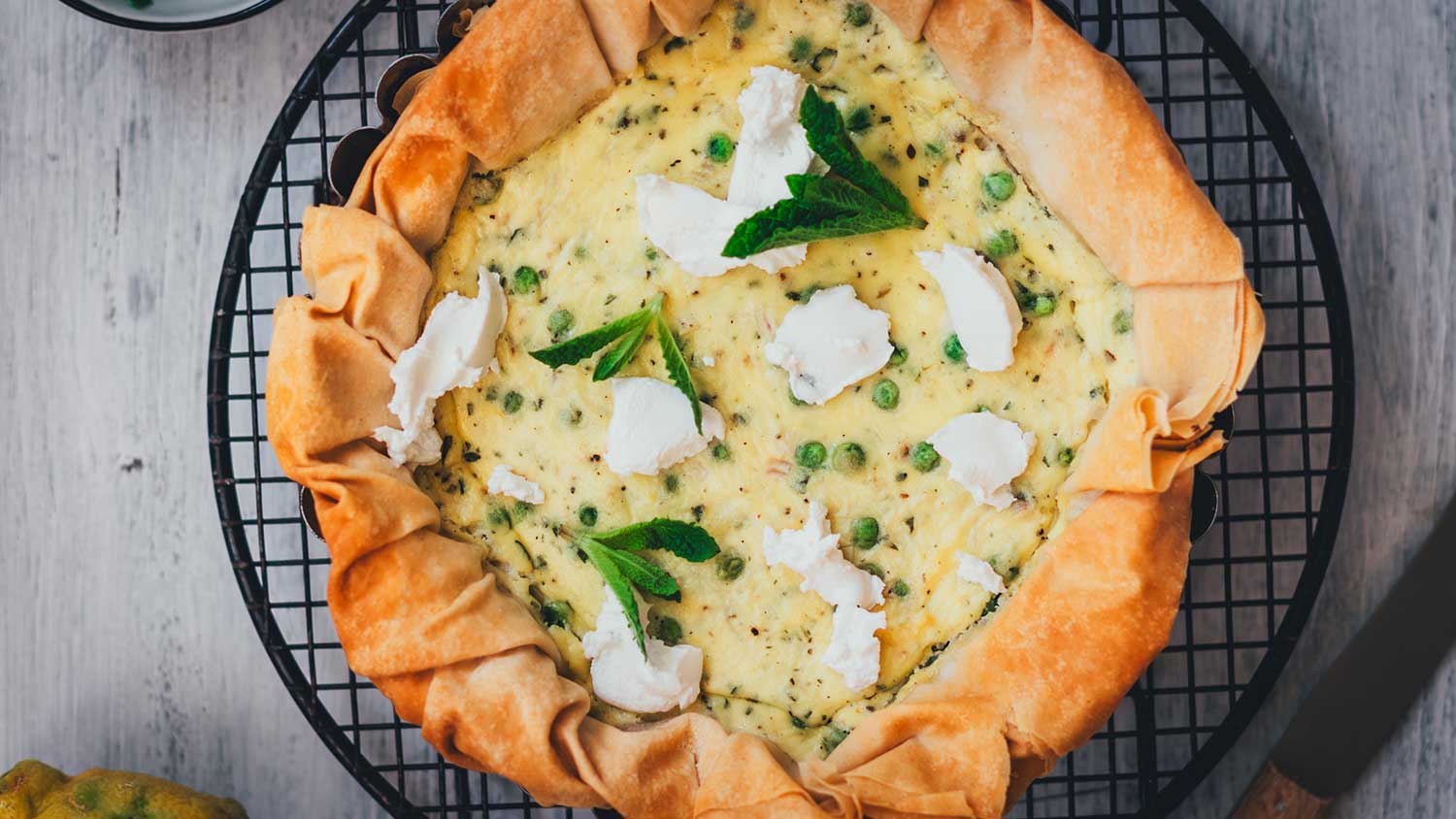 Erbsen-Ricotta-Quiche mit Filoteig in einer schwarzen Tarteform auf einem runden Auskühlgitter, garniert mit Tupfen Ziegenfrischkäse und frischen Minzeblättern, von oben fotografiert.