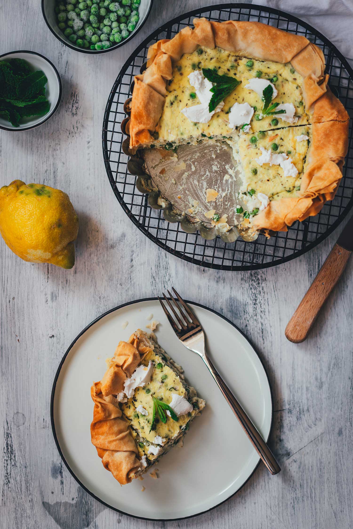 Angeschnittene Erbsen-Ricotta-Quiche mit Filoteig, ein serviertes Stück liegt mit Gabel auf einem hellen Teller, daneben TK-Erbsen, Minze und eine Zitrone.
