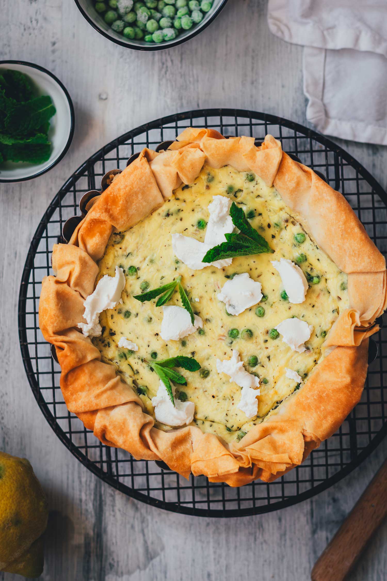 Vegetarische Erbsen-Ricotta-Quiche mit knusprigem Filoteig-Rand, garniert mit Ziegenfrischkäse und Minze, von oben auf hellem Holzuntergrund mit TK-Erbsen und Zitrone.