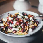 Angerichteter gegrillter Zucchini-Salat mit Oliven und Ziegenfrischkäse auf hellem Teller, im Hintergrund die Küche